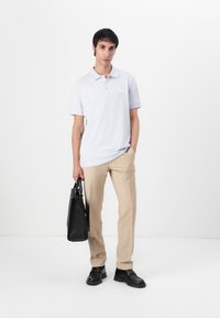 Svijetloplena polo majica s dugmetima, khaki hlače i crna kožna torba. Model je postavljen naspram bijele pozadine.