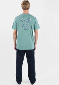 T-shirt manches courtes couleur bleu sarcelle clair en coton, avec un motif floral coloré dans le dos. Associé à un pantalon bleu marine foncé.