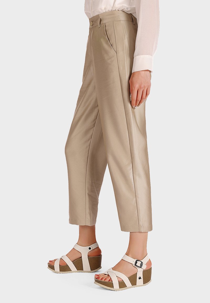 Pantaloni beige a vita alta con una texture liscia e pieghe frontali, abbinati a sandali con tacco a forma di cuneo bianchi e con una base simile al sughero.