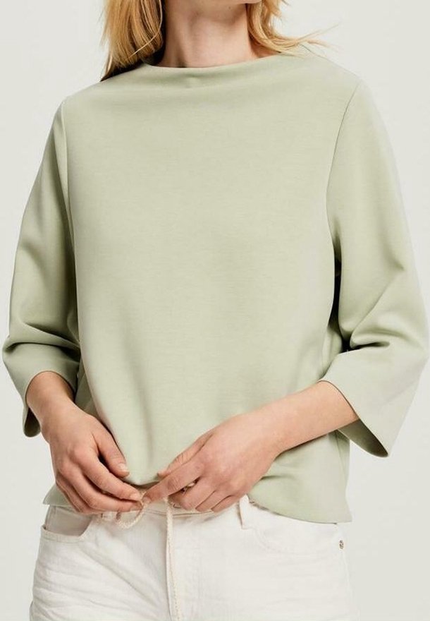 Femme portant un haut vert clair à manches longues avec de larges poignets, ajustant l'ourlet, associé à un pantalon blanc sur un fond neutre.