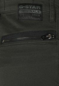 Tummanvihreä denimi, jossa on musta vetoketjupussi ja musta suorakulmainen laikku, jossa lukee "G-STAR ORIGINALS RAW DENIM."