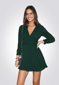 Vestido verde con puntos, escote en V, mangas largas, cuerpo fruncido y dobladillo con volantes. Combinado con un pequeño bolso negro.