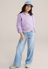 Fille portant une casquette de baseball bleu clair, un t-shirt à manches longues lavande, un jean large bleu clair et des baskets beige, debout devant un fond beige.