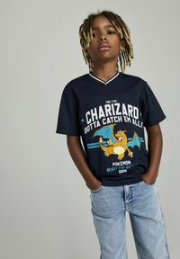 Camiseta gráfica azul marino con Charizard, texto "Tipo Fuego" y "¡Consíguelos Todos!", con mangas cortas y un escote de color contrastante.