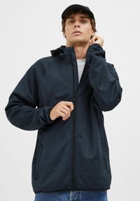 Jūrinė zip-up striukė su gobtuvu, turinti reguliuojamą apykaklę, minkštą tekstūrą, dvi šonines kišenes ir reglaninius rankoves. Derinama su mėlynomis džinsinėmis kelnėmis.