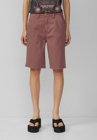 Roze spijkerhortens met een rechte snit, tot op de knie, voorzien van riemlussen en een knoopsluiting. Draagt met zwarte platform sandalen.