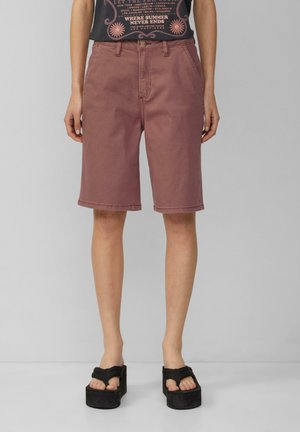Shorts en denim rose avec une coupe droite, arrivant au genou, dotés de passants de ceinture et d'une fermeture à bouton. Portés avec des sandales à plateforme noires.