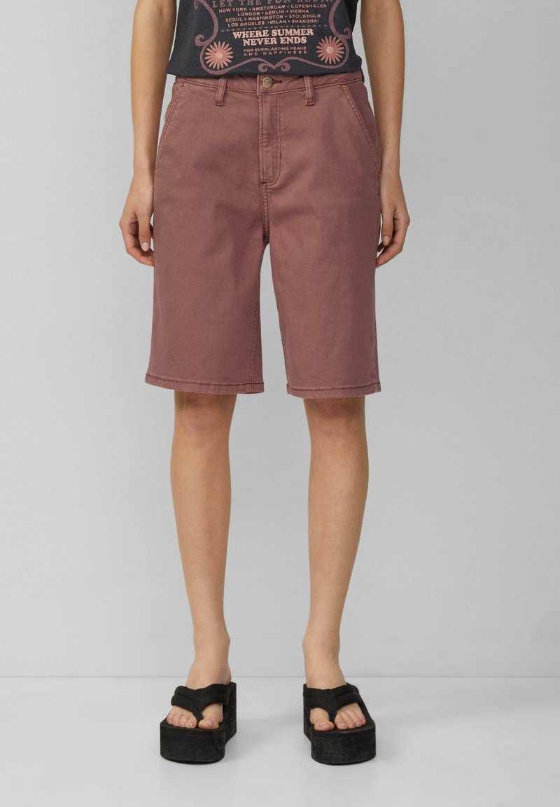 Roze spijkerhortens met een rechte snit, tot op de knie, voorzien van riemlussen en een knoopsluiting. Draagt met zwarte platform sandalen.