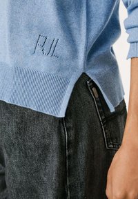 Šviesiai mėlyna megzta savanorės su raukšlėmis ir šoninėmis skeltimis, papuošta išsiuvinėtu "PUL" užrašu. Dėvima su tamsiai mėlynomis džinsinėmis kelnėmis.