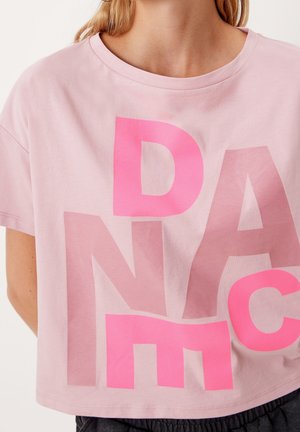 Persoon die een lichtroze T-shirt met korte mouwen draagt met grote overlappende letters in donkerder en helderder roze.