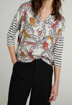 Blouse blanche à manches longues ornée d'un imprimé animal coloré avec des motifs de léopard et de lion, associée à un pantalon noir pour un contraste.