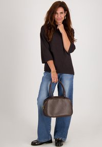 Bruine leren handtas met een ronde vorm, dubbele hengsels en een rits. Draagt door een persoon in een zwarte top en blauwe jeans. Eenvoudig ontwerp.