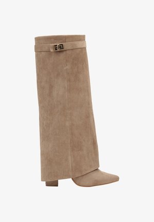 Botte beige en daim montant jusqu’au genou, avec un talon bloc, un bout pointu et une bride décorative avec une boucle dorée près du haut.