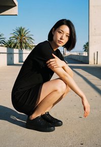 Tânără cu păr negru scurt stă în poziție de ghemuit pe o potecă exterioară de beton însorită, purtând o rochie neagră și adidași negri.