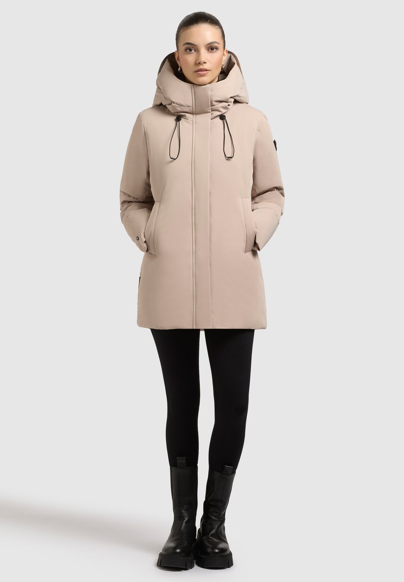 Beige hooded puffer jacket with a slim fit, zip closure, drawstring accents, and side pockets, paired with black leggings and chunky boots.