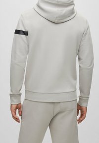 Ljust grå hoodie med en texturerad yta, som har ett svart rand på vänster ärm, ribbade muddar och en mjuk, avslappnad passform.