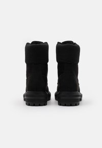 Par de botas de invierno de ante negro con suelas de goma gruesas y puños de felpa doblados, vistas desde la parte de atrás sobre un fondo blanco.
