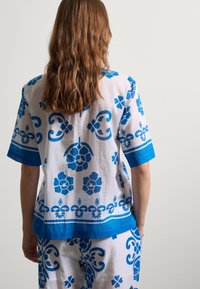 Witte shirt met blauwe bloemen- en spiraalpatronen, korte mouwen, blauwe afwerking en een losse pasvorm. Draagt bijpassende broek met blauw patroon.