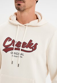 Kremowa bluza z kapturem z czerwonym nadrukiem graficznym, na którym widnieje napis "Creeks ORIGINAL Since 1974", z trokami i kieszenią kangurką.