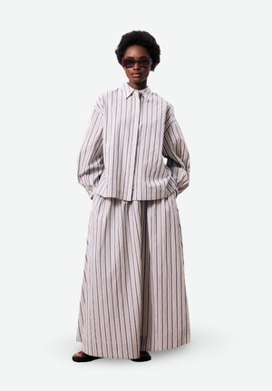 Persoon draagt een oversized, verticaal gestreept overhemd met knopen en brede, bijpassende gestreepte broek met donkere sandalen en zonnebril.