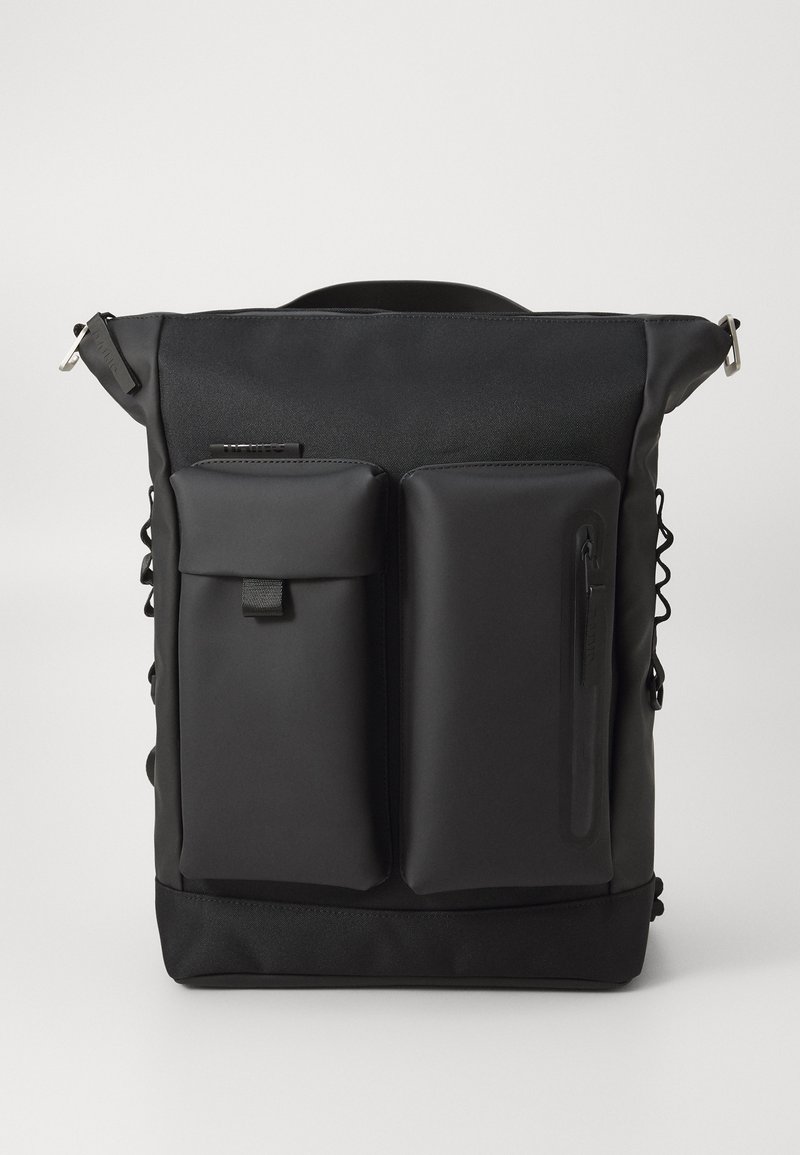 Mochila preta com múltiplos compartimentos, textura suave, fechos de zip e alças ajustáveis. Possui ainda bolsos laterais adicionais para acessórios.