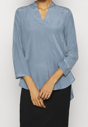 Žena nosící světle modrou blůzu s dlouhým rukávem a výstřihem do V a černou sukní, ruce sepnuté v pase, stojící před jednobarevným pozadím.