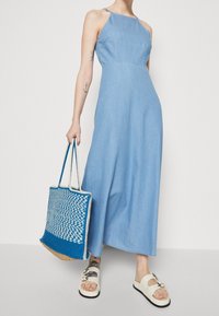 Vaaleansininen denim-maksimekko, jossa on istuva yläosa ja leveä hame. Asusteena sininen kudottu olkalaukku ja valkoiset litteät sandaalit.