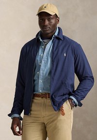 Mann mit beigefarbener Kappe, dunkelblauer Jacke über blauem Jeanshemd und gestreiftem Oberteil, beige Hose mit geflochtenem Gürtel, Hand in der Tasche vor einfarbigem Hintergrund.