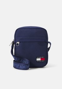 Bolso cruzado de tela azul marino con cierre de cremallera, que cuenta con un bolsillo frontal y una correa de marca con "TOMMY JEANS" en letras azules.