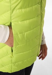 Limegrön pufferväst med kviltat mönster och en dragkedjeficka, med glänsande material och hög krage, bärs över en vit långärmad skjorta.