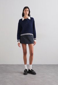 Pulover din lână bleumarin cu model tricotat, cu guler alb și tiv cu dungi, asortat cu o fustă mini neagră. Modelul poartă pantofi negri și șosete albe.