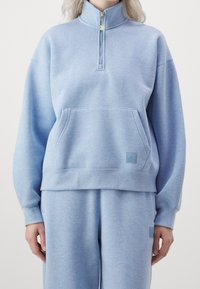 Hellblaues Sweatshirt mit hohem Kragen, halbem Reißverschluss, Fronttasche und dezentem Logo-Patch. Weicher Stoff mit lockerer Passform und gerippten Manschetten.