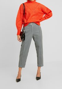 Osoba oblečená v jasně oranžovém pleteném svetru, černobílých kalhotách s houndstooth vzorem, černých lodičkách a držící černou kabelku.