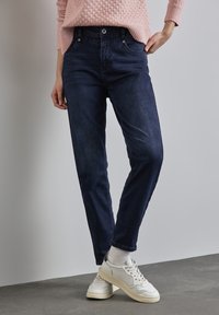 Mörkblå denimbyxor med klassisk passform, fem fickor och knäppning. Matchat med vita sneakers och en ljusrosa tröja.