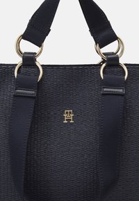 Gros plan sur un sac cabas tissé noir avec de larges bretelles noires attachées par des anneaux dorés et un petit logo doré aux lettres entrelacées.