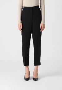 Pantalones negros de corte ajustado, con una cintura alta y longitud recortada. Llevados con tacones altos y brillantes negros. Textura de tela suave.