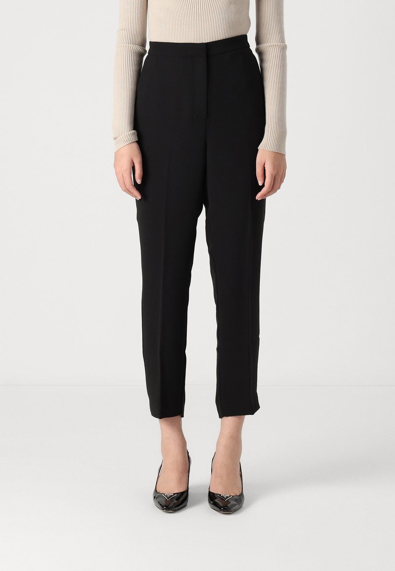Pantalones negros de corte ajustado, con una cintura alta y longitud recortada. Llevados con tacones altos y brillantes negros. Textura de tela suave.