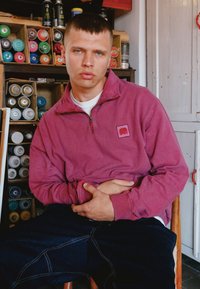 Ružový flísový mikina na zips so štvorcovou nášivkou, spojená s tmavomodrými džínsami. Pozadie obsahuje plechovky so sprejom.