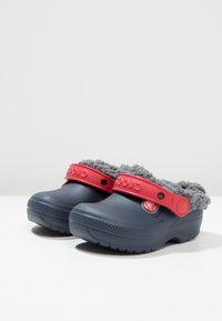 Granatowe chodaki z miękkim szarym futerkiem w środku oraz jaskrawoczerwonym regulowanym paskiem. Teksturowana podeszwa zapewnia przyczepność. Detal logo na pasku.