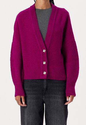 Frau trägt eine magentafarbene, geknöpfte Strickjacke mit gerippten Ärmeln über einem grauen Top und schwarzen Jeans, stehend vor einem einfarbigen Hintergrund.