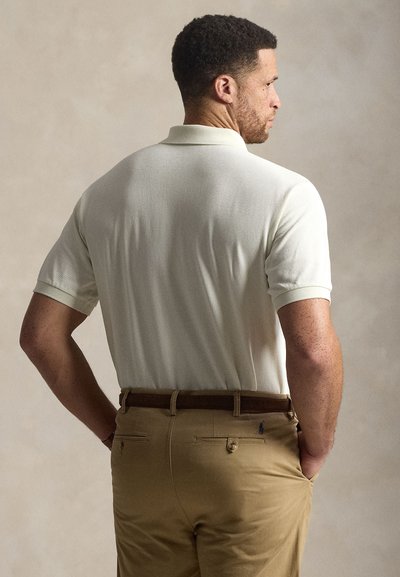 Homme aux cheveux courts et bouclés, portant un polo blanc rentré dans un pantalon beige avec une ceinture marron, debout de dos, les mains dans les poches.