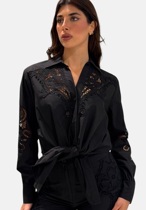 Donna che indossa una maglia nera a maniche lunghe con ritagli decorativi in pizzo su spalle e maniche, legata in vita, che guarda leggermente a destra.