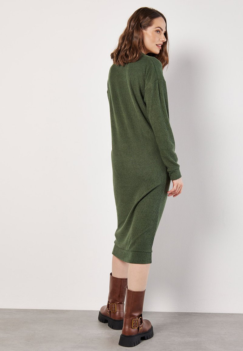 Grünes geripptes Strickkleid mit langen Ärmeln, seitlichen Schlitzen und einer Midi-Länge, kombiniert mit braunen Lederstiefeln mit Schnallen.