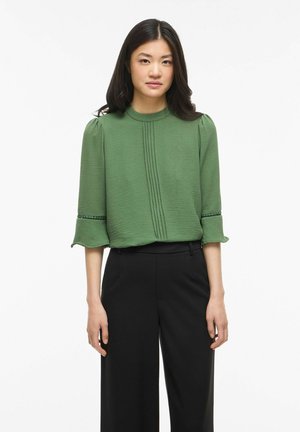 Femme portant un chemisier vert texturé avec des plis et des détails de perles sur les manches, associé à un pantalon noir large taille haute, debout sur fond blanc.