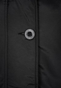 Detail černé péřové bundy s lesklým černým knoflíkem a texturovanou látkou s viditelným švem, vystavené na jednolitém pozadí.