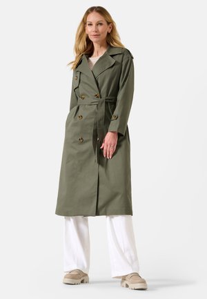 Trench-coat vert olive avec une ceinture, design croisé à double boutonnage et boutons dorés ; porté sur un pantalon large blanc et des chaussures beiges.