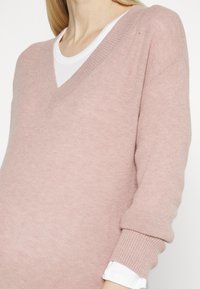 Sweater rosa em V, feito de um tecido de malha suave, com punhos canelados e uma camisa branca em camadas por baixo. Textura lisa com padrão subtil.