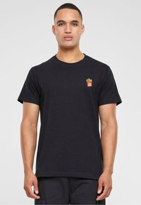 Tricou negru din bumbac, cu un mic design brodat în galben și roșu reprezentând cartofi prăjiți, situat în zona stângă a pieptului. Mâneci scurte.