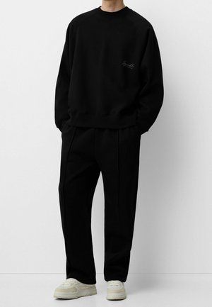 Persona con sudadera negra y pantalones negros con zapatillas blancas, de pie frente a un fondo blanco liso.