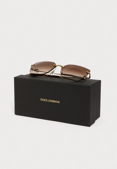 Hnědé bezrámové sluneční brýle se zlatými akcenty ležící na černé krabičce značky Dolce & Gabbana na bílém pozadí.